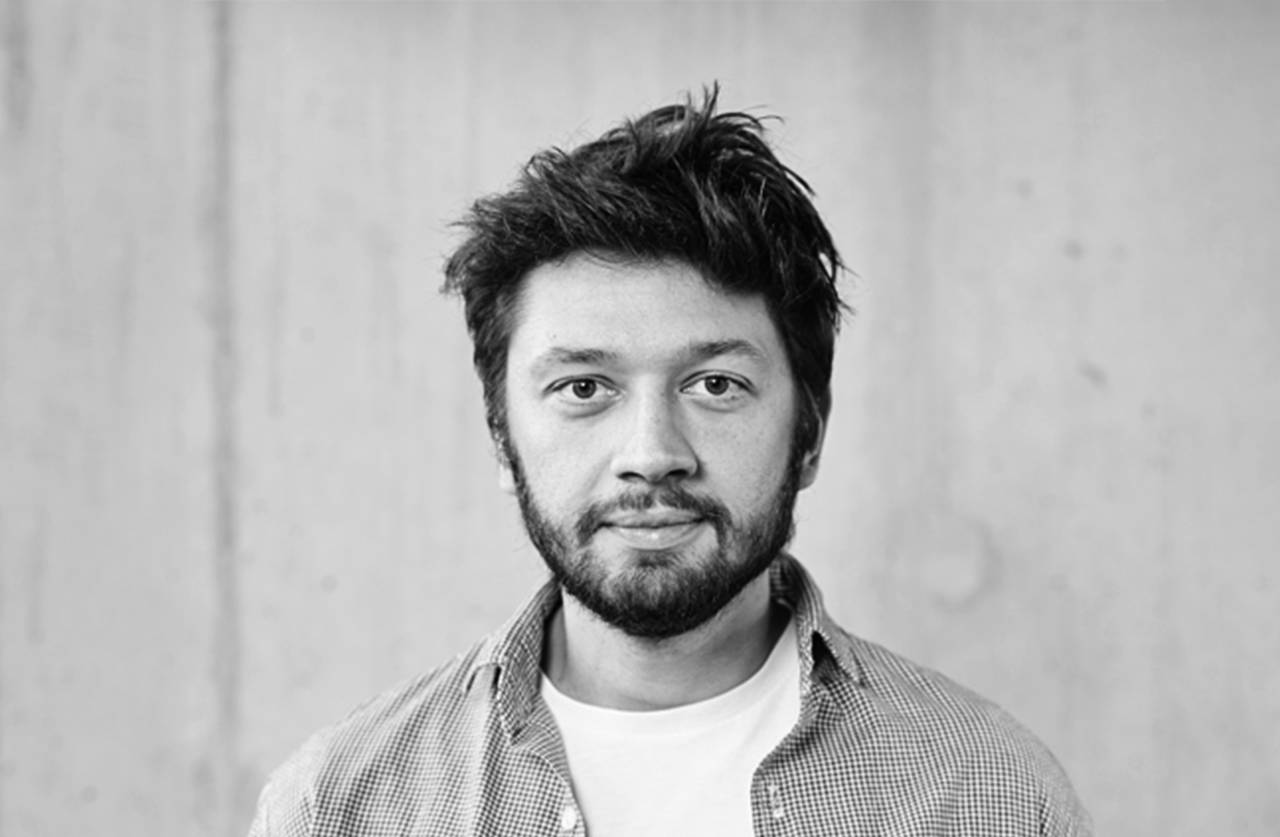 A man with a beard and tousled hair wearing a checkered shirt over a white t-shirt looks directly at the camera. The background is plain and blurred, creating a simple portrait in black and white.