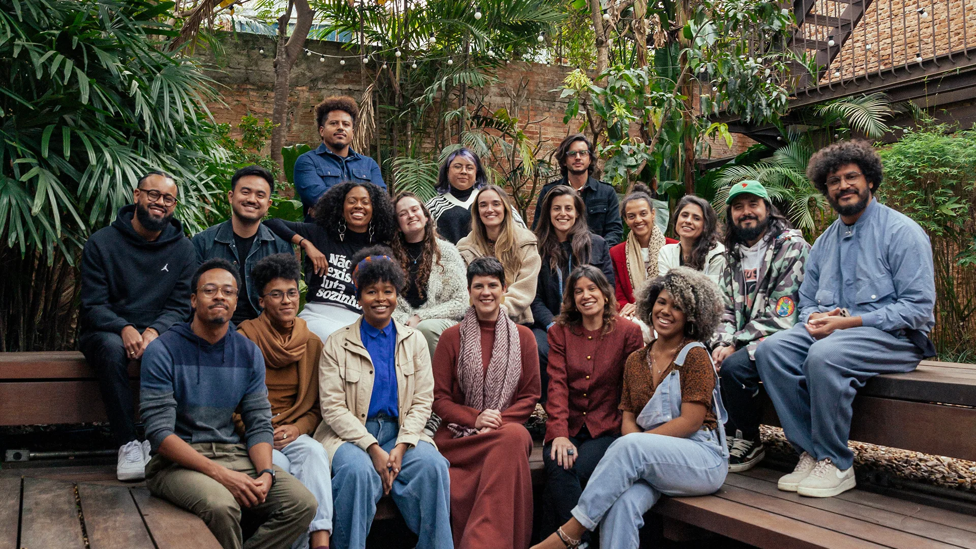 A group of 20 people of diverse backgrounds sit and stand together on wooden benches in an outdoor setting with lush greenery, smiling at the camera.