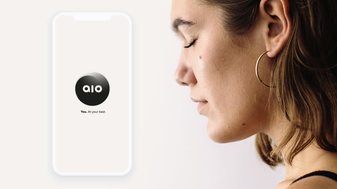 A woman with short brown hair and a gold hoop earring faces right, eyes closed, beside a glowing smartphone displaying the logo aio and the text You. At your best. on a white background.