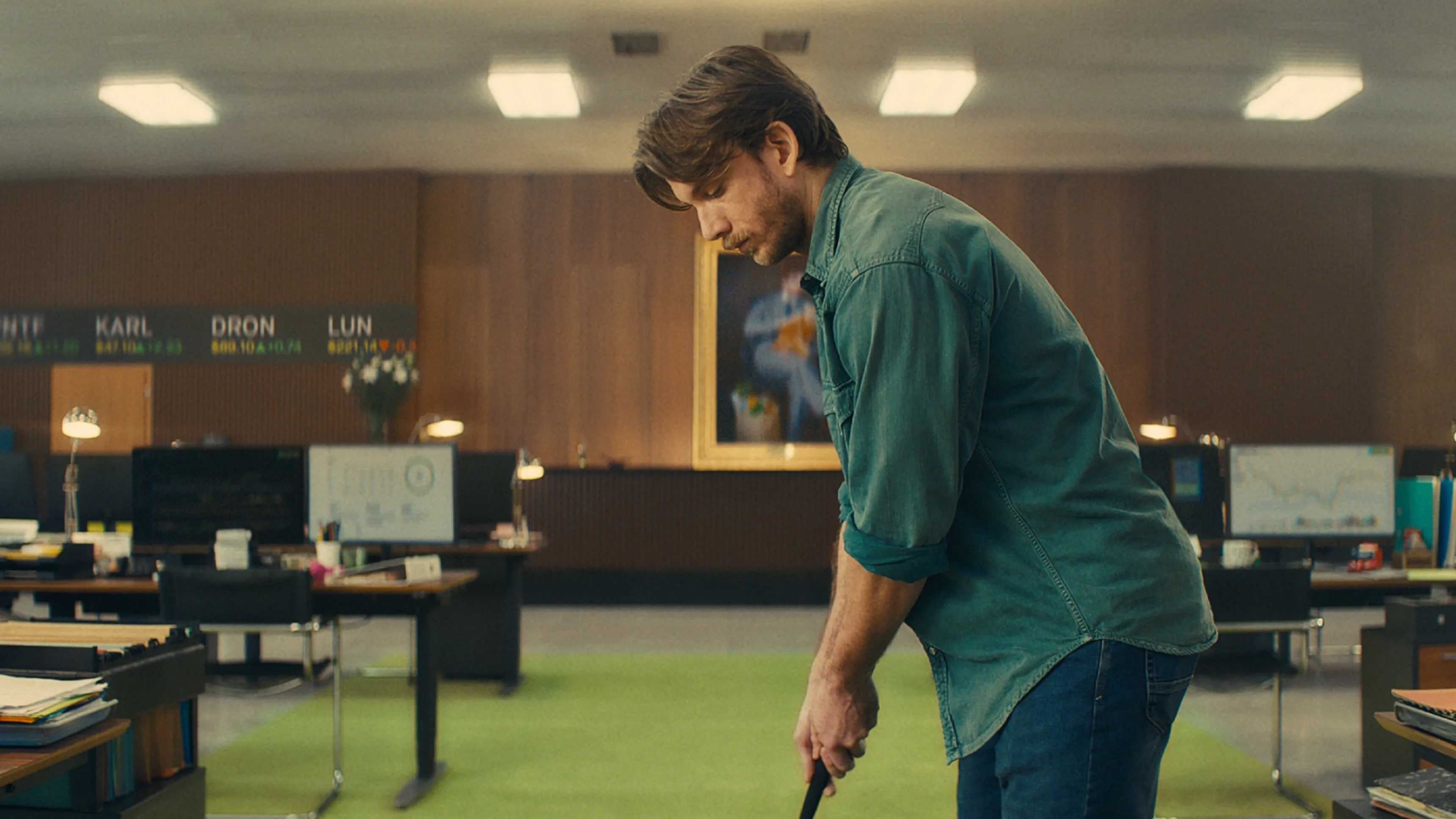 A man in a green shirt plays mini-golf in the middle of an office with desks, computers, and charts on the walls. The office has wood paneling and bright ceiling lights.
