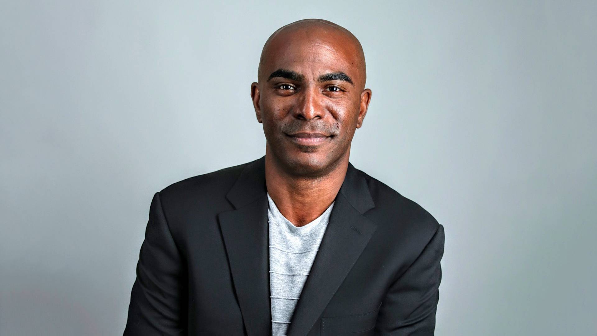 A man with a shaved head wearing a black blazer over a light gray shirt smiles slightly while sitting against a plain gray background.