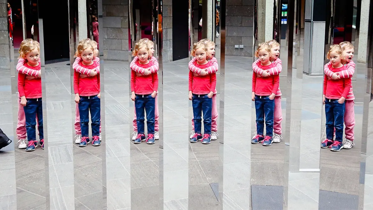 Two young children stand together indoors, one hugging the other from behind. Their reflections appear multiple times in tall, vertical mirrors, creating the illusion of many identical pairs.