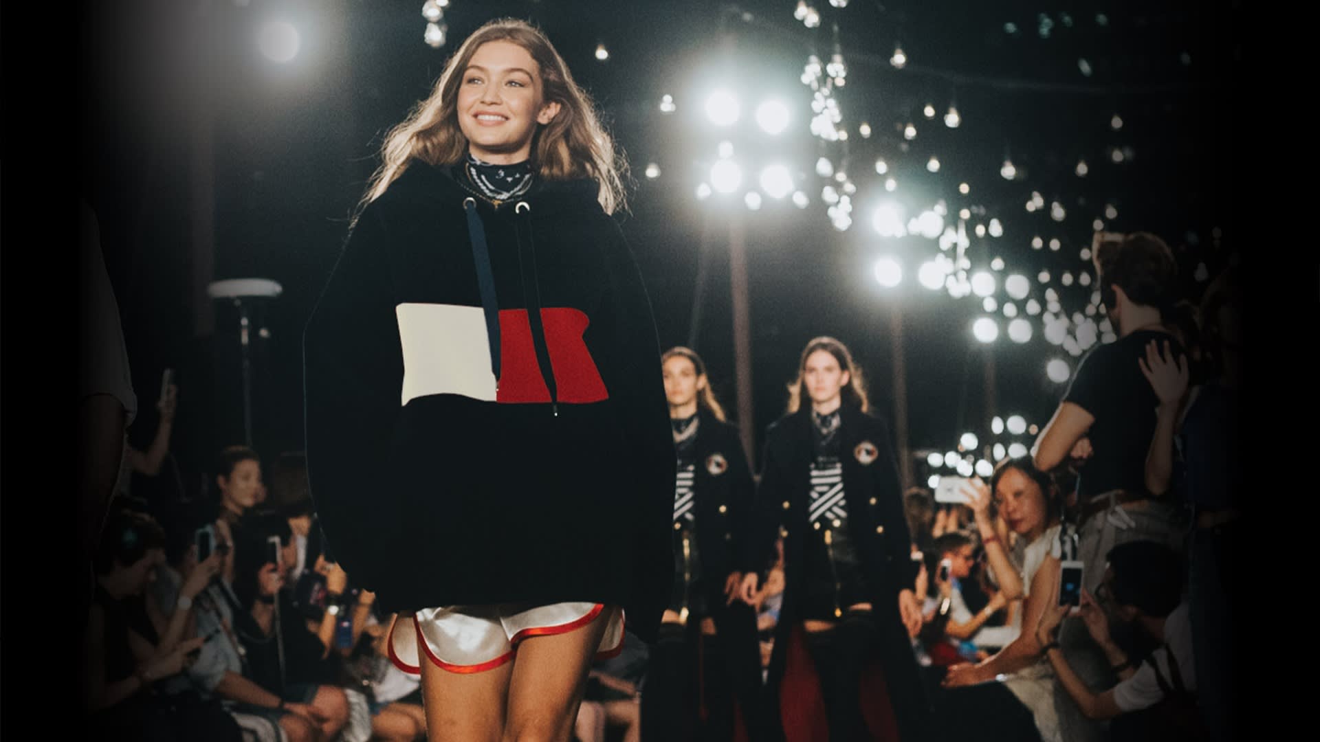 A smiling model walks the runway wearing a black hoodie with a bold red and white design and white shorts, surrounded by glowing lights and an audience, with other models following behind.