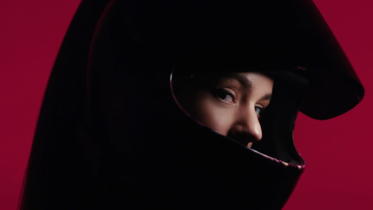 A person wearing a black helmet looks intently through the visor. The background is a solid, vibrant red, highlighting the dramatic contrast with the helmet and the focused expression on their face.