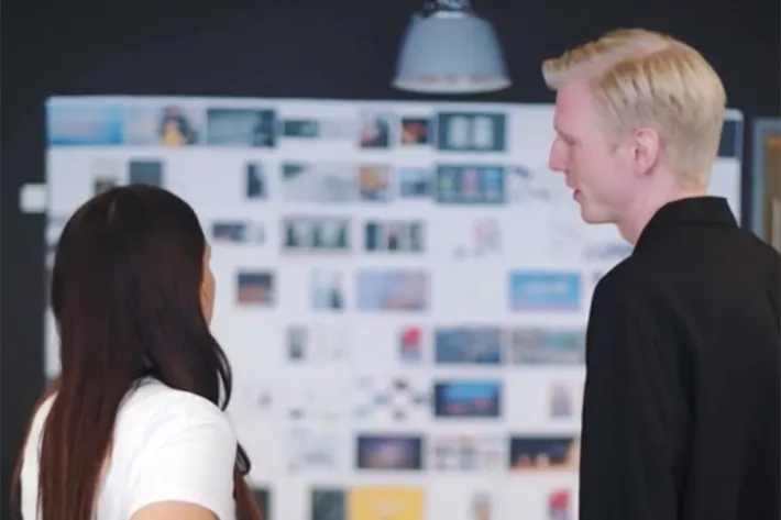 Two people, one with long dark hair and one with short blond hair, stand in front of a wall covered in various photos and images, appearing to discuss or review them.