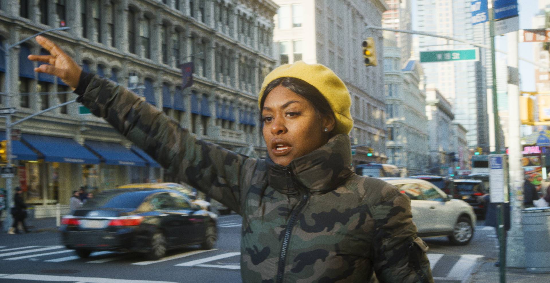 A woman in a camouflage jacket and yellow beret stands on a busy city street, raising her arm as if hailing a taxi. Cars and tall buildings surround her, and street signs are visible in the background.