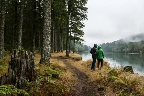 Two people in hooded jackets stand on a forest path beside a calm lake, surrounded by tall trees and misty, overcast weather. The ground is covered in grass and fallen branches.