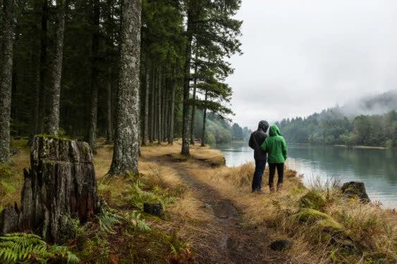 Two people in hooded jackets stand on a forest path beside a calm lake, surrounded by tall trees and misty, overcast weather. The ground is covered in grass and fallen branches.