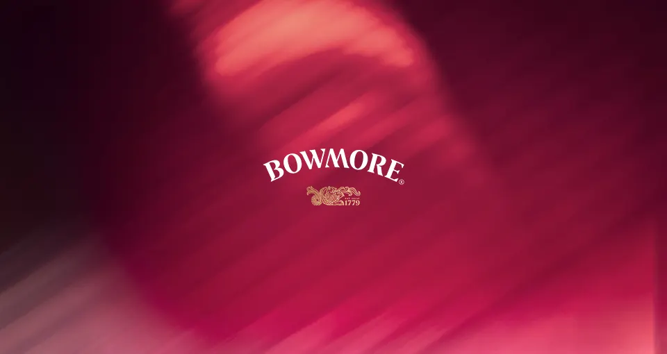 The image features the word BOWMORE in white uppercase letters, with a golden ornate design and the year 1779 below, set against a blurred background of deep pink and red hues.