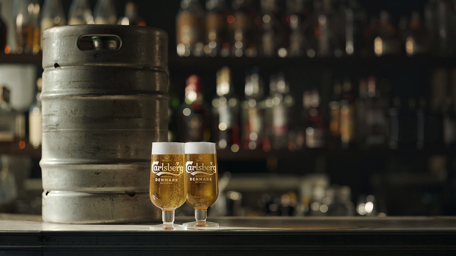 Two glasses of Carlsberg beer with foam on top sit on a bar counter next to a metal keg, with shelves of assorted liquor bottles blurred in the background.