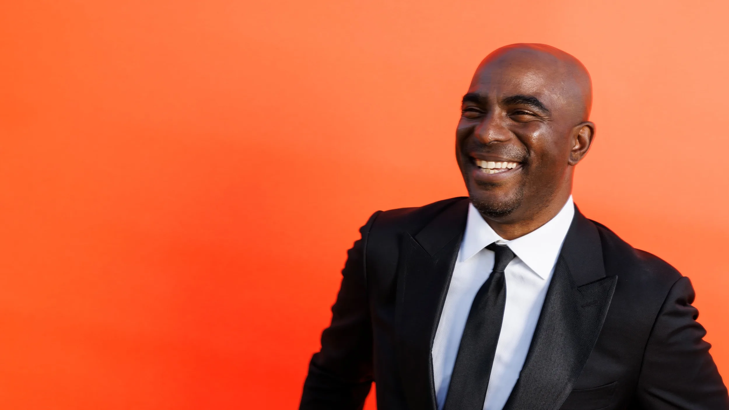 A man in a black suit and tie smiles warmly while standing in front of a bright orange background.