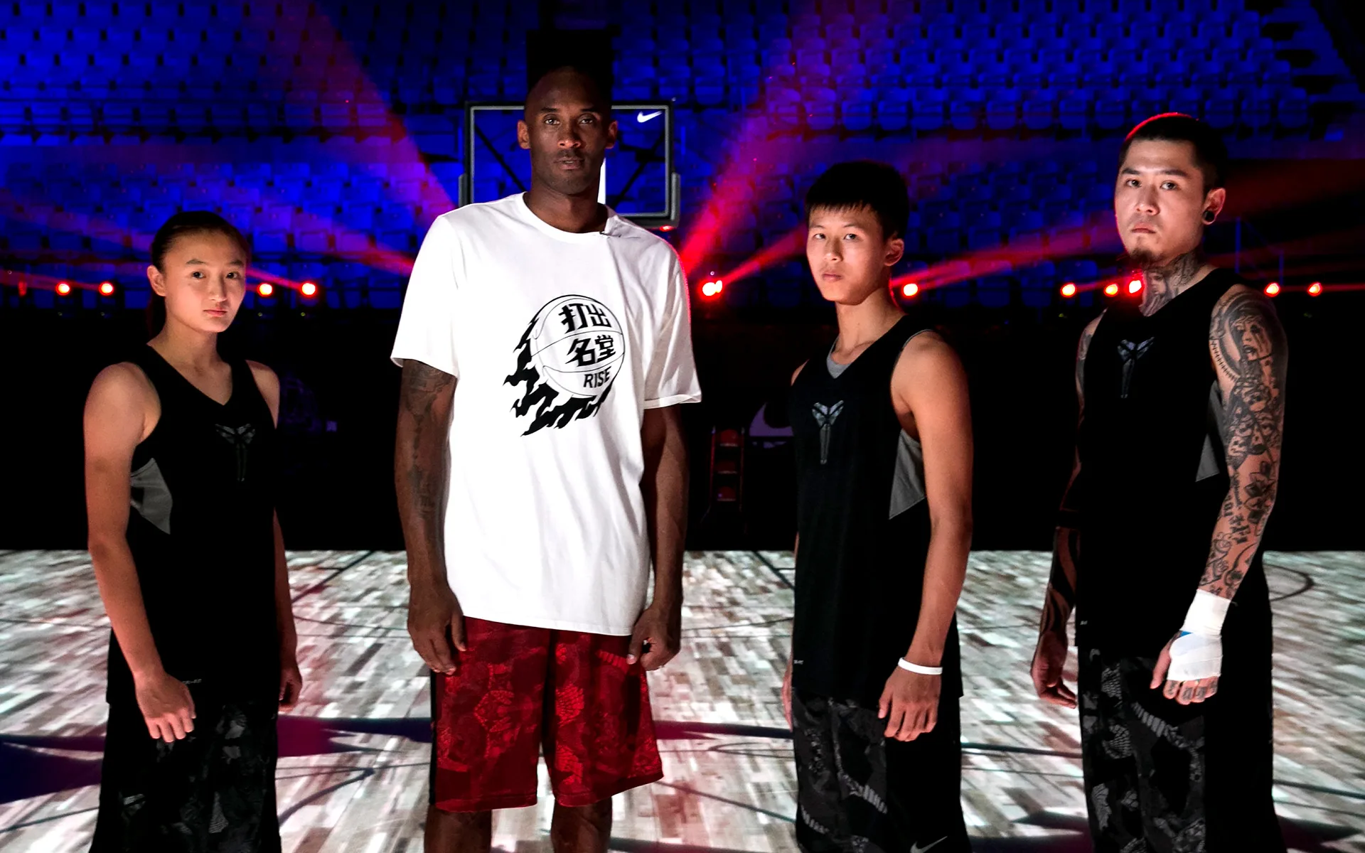 A tall man in a white shirt and red shorts stands on a basketball court with three younger people in matching black athletic outfits, under dramatic lighting in an indoor arena.