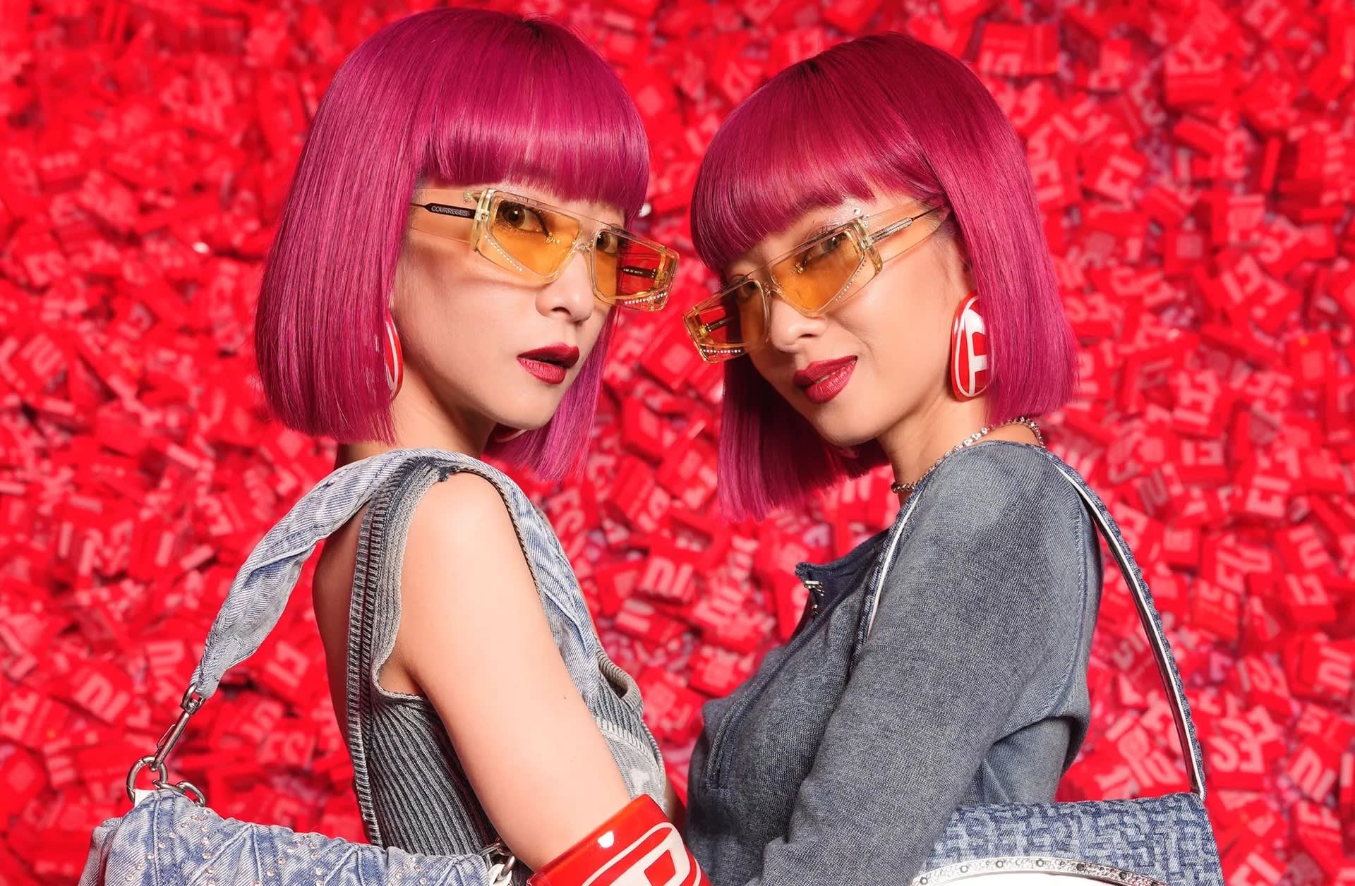 Two women with matching vibrant magenta bob hairstyles, wearing orange-tinted sunglasses, bold red lipstick, gray outfits, and large statement earrings, stand closely together in front of a background covered in red items.