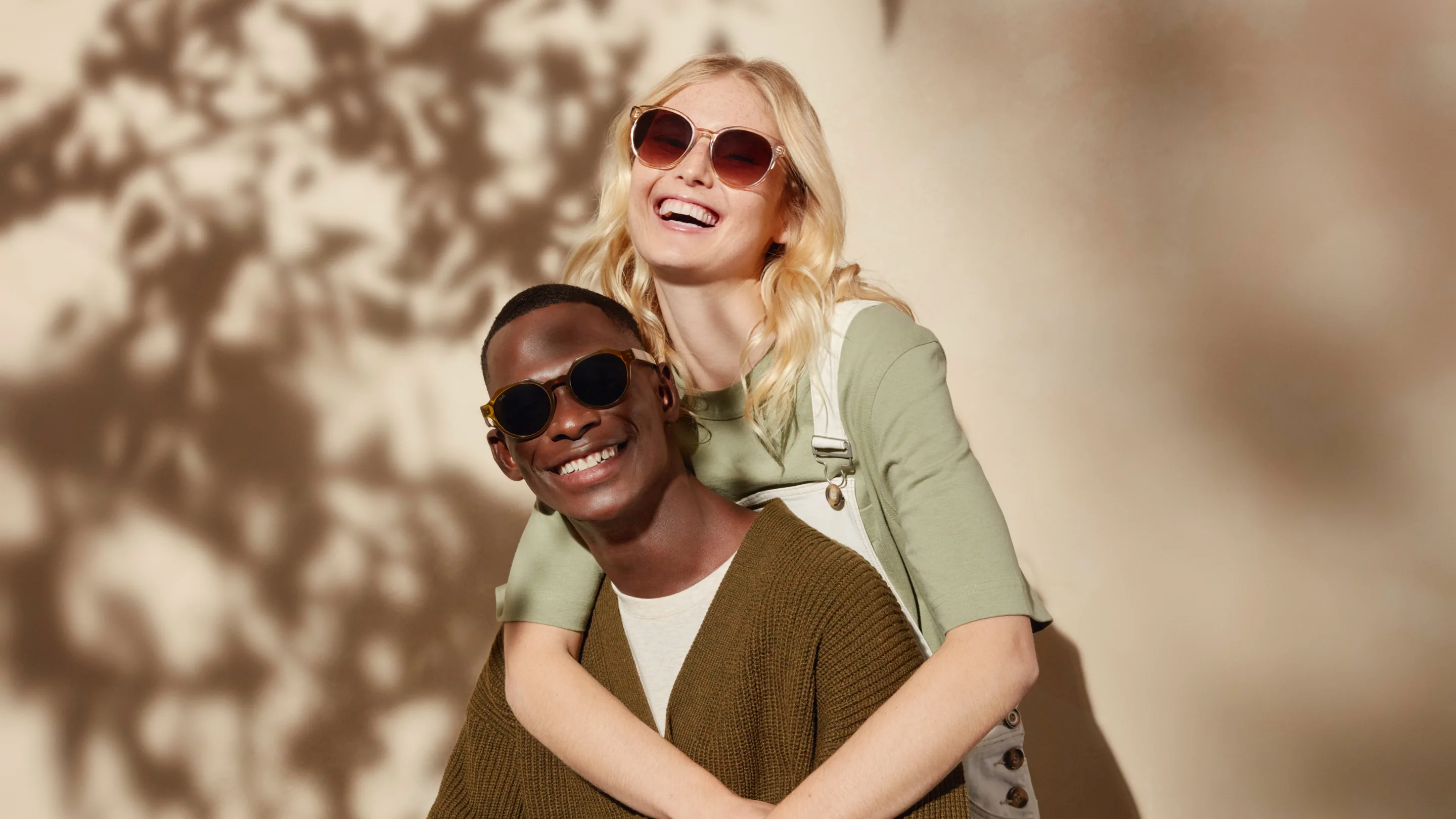 Two people wearing sunglasses smile and pose together against a sunlit beige wall with leafy shadows. One stands behind and hugs the other from behind, both looking happy and relaxed.