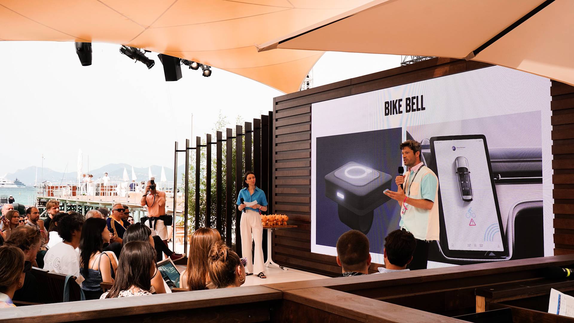 A man speaks to an audience at an outdoor event, standing next to a large screen displaying the words “BIKE BELL” and a phone image, while another person stands nearby. The setting overlooks water and boats in the background.