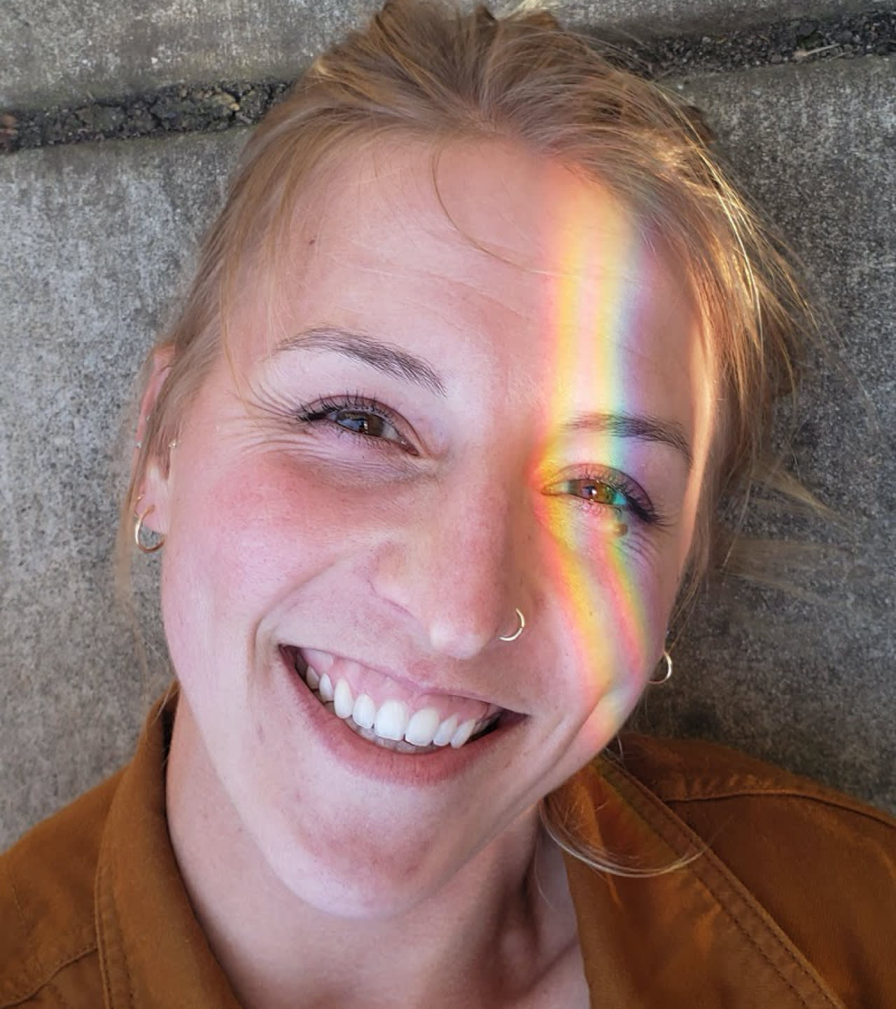 A smiling woman with light brown hair lies on concrete, sunlight casting a vivid rainbow across her face from forehead to cheek. She wears a nose ring, small earrings, and a brown jacket.