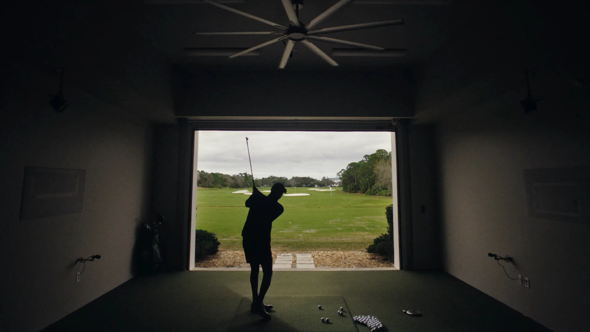 A person swings a golf club inside a darkened room with an open doorway, facing a green golf course under a cloudy sky. Golf balls and tees are scattered on the indoor turf near their feet.