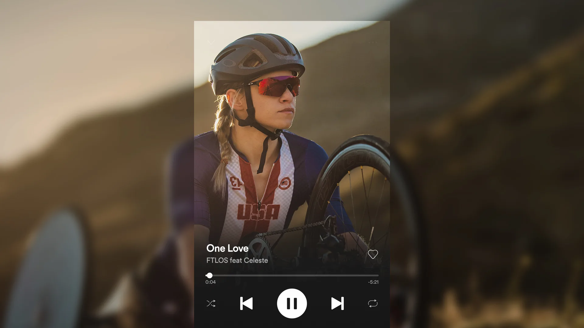 A female cyclist in a USA uniform and sunglasses is outdoors, holding a handcycle. A music player overlay shows One Love by FTLOS feat Celeste is playing.