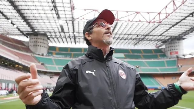 A man wearing glasses, a cap, and a black AC Milan jacket stands in an empty stadium, looking up with his arms slightly raised, under a cloudy sky and the stadium’s open roof structure.