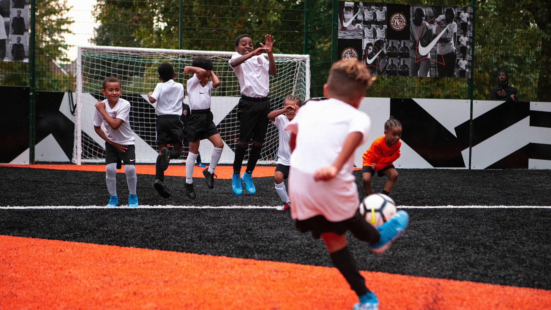 A group of children playing soccer on an outdoor field; one child prepares to kick the ball while others in white uniforms jump in front of the goal to block it. The field has bold black and orange markings.