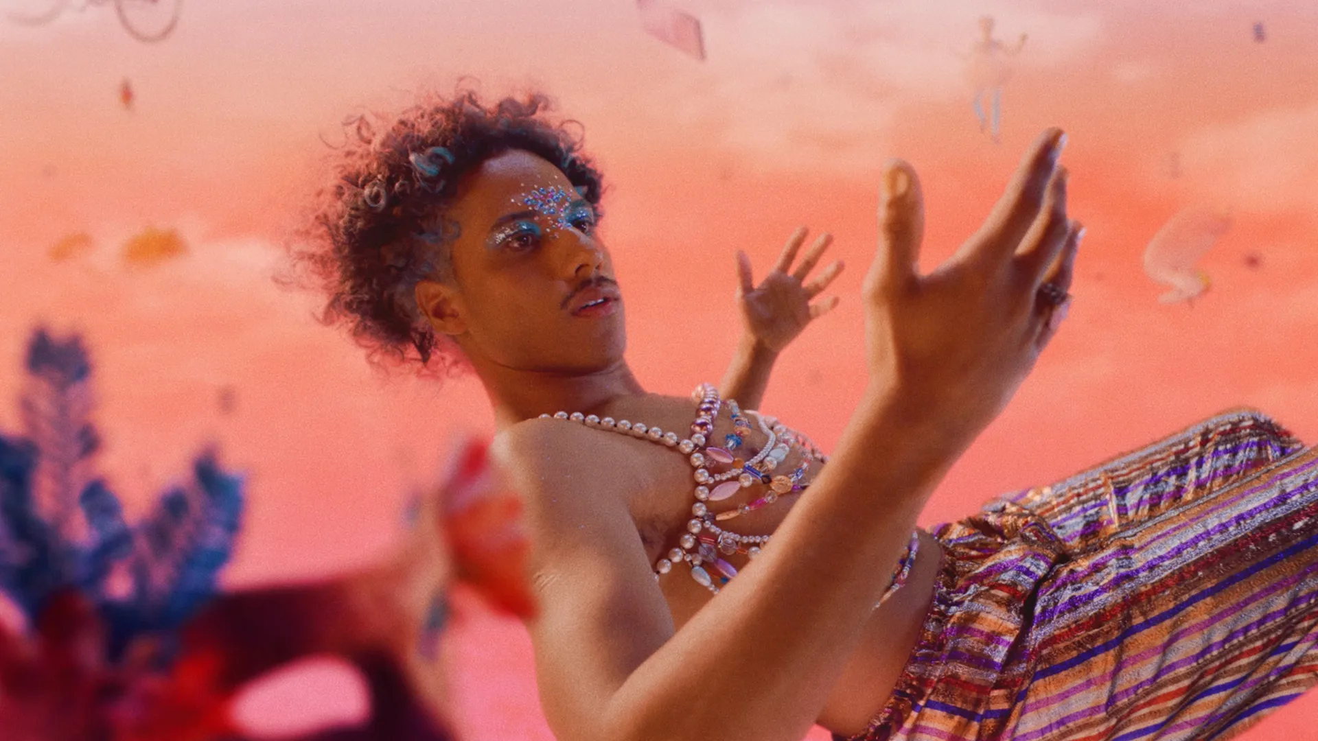 A person with curly hair and bright blue rhinestones around their eyes wears pearl jewelry and a sparkly striped outfit, floating against a surreal pink and orange sky with abstract shapes in the background.