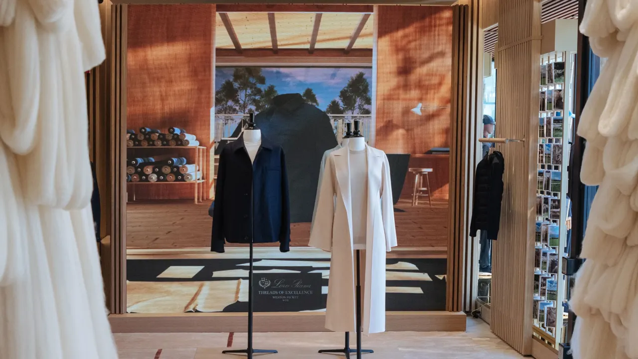 Two mannequins display a black coat and a white coat in a stylish boutique, with fabric rolls on shelves and a large scenic photo backdrop behind them featuring a sunny room with trees visible through windows.