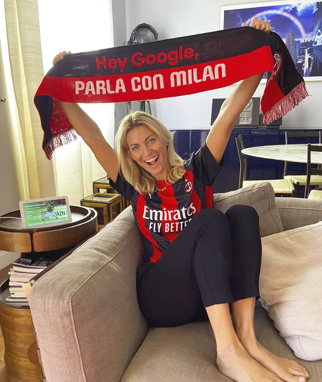 A smiling woman in an AC Milan jersey sits on a couch, holding up a red and black scarf that says Hey Google, PARLA CON MILAN. A smart display is visible on the table beside her.
