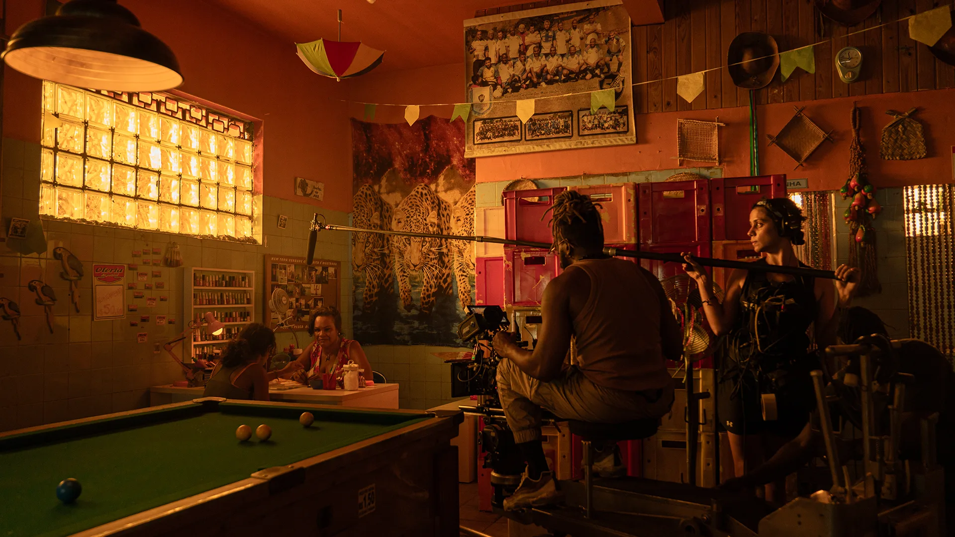 A film crew with camera and boom mic records in a warmly lit café decorated with photos and paintings, while people sit at tables and a green pool table is in the foreground.