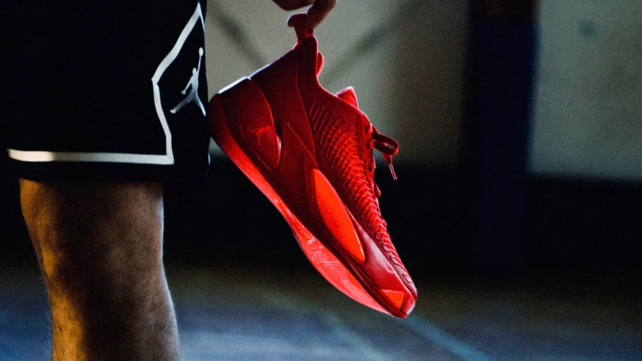 A person wearing black shorts holds a bright red athletic sneaker by the heel, with the shoe dangling next to their leg against a dim, blurred indoor background.
