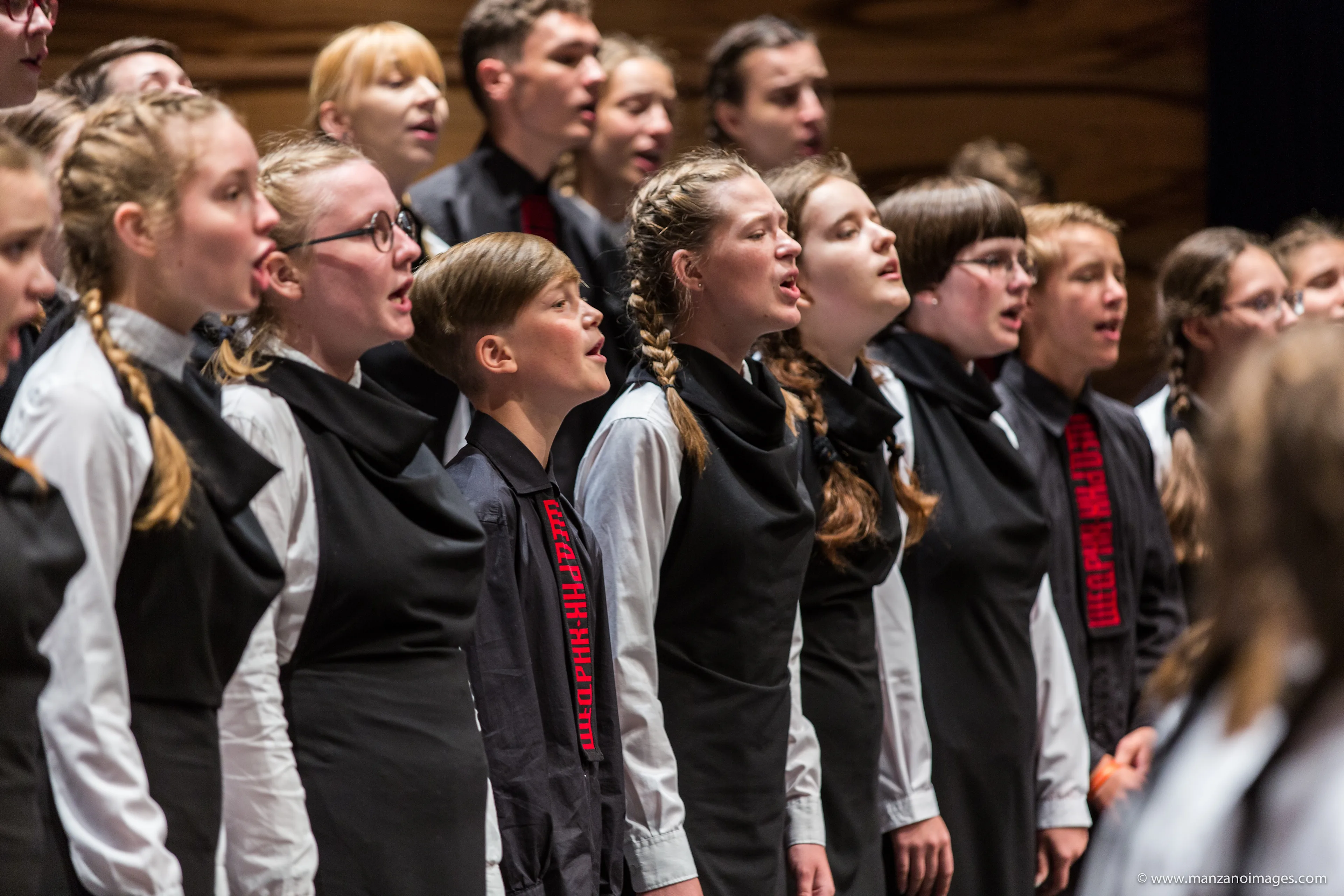 A group of young choir singers, dressed in matching black and white outfits, sing passionately together on stage. They stand in rows, focused and engaged in their performance.