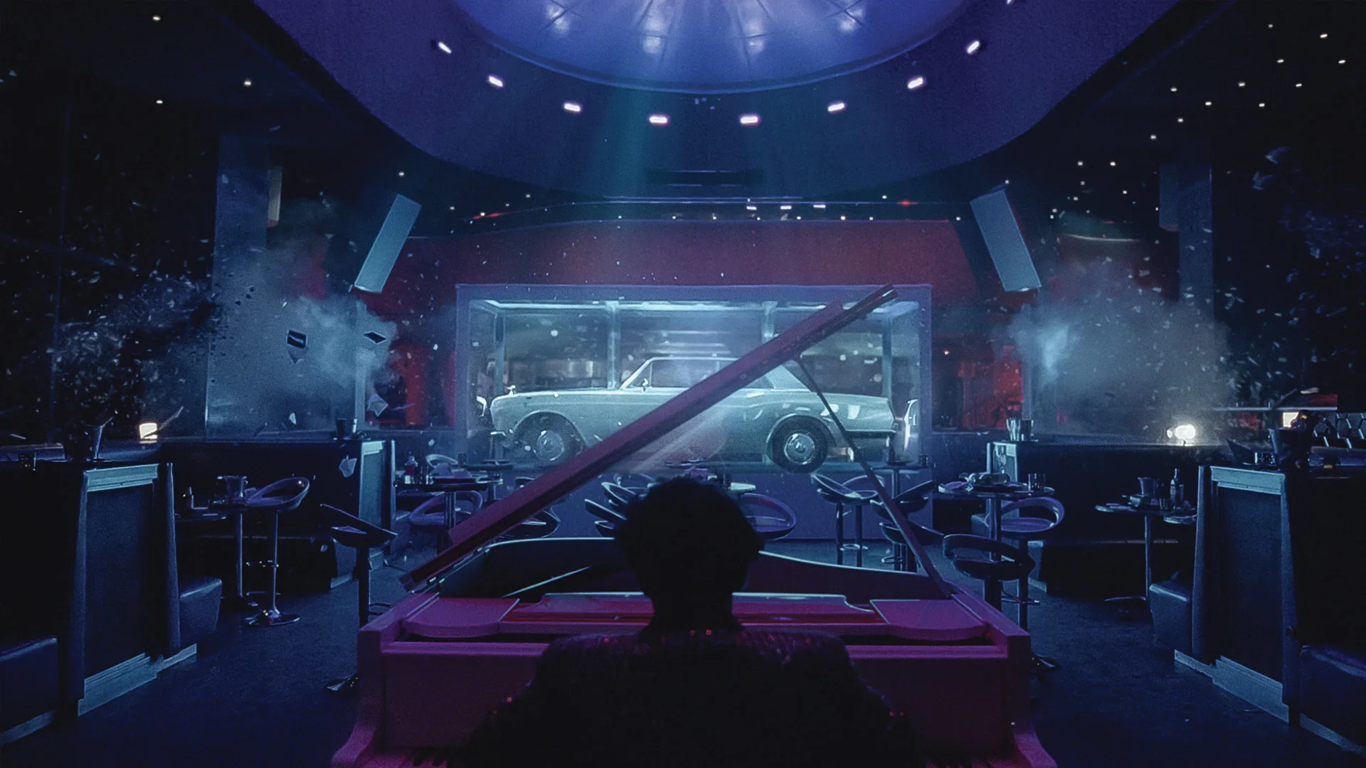 A person plays a grand piano in an empty, dimly lit nightclub. In the background, a vintage car is displayed inside a glass case, illuminated by blue and purple lights. Tables and chairs are scattered around the room.