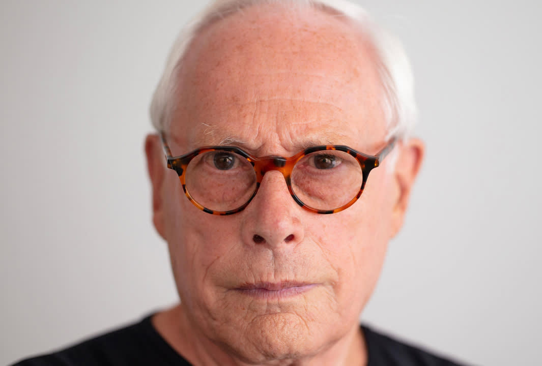 An older man with short white hair wearing round, tortoiseshell glasses looks seriously at the camera. He is dressed in a dark shirt and stands against a plain light background.