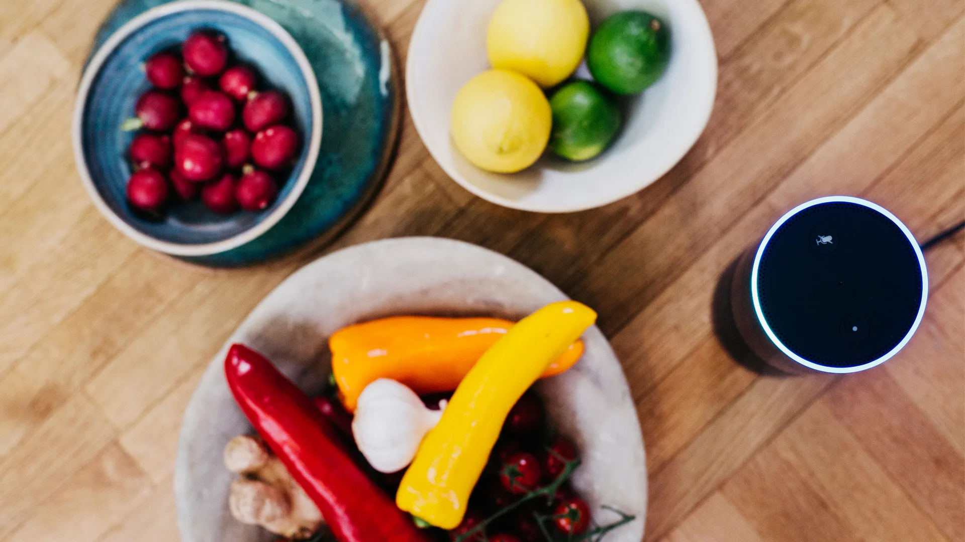 Three bowls on a wooden table: one with red radishes, one with lemons and limes, and another with colorful peppers, garlic, ginger, and tomatoes. An Amazon Echo smart speaker is nearby.