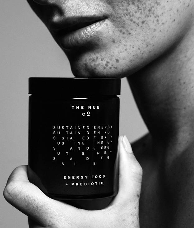 A person with freckles holds a dark jar labeled THE NUE CO. SUSTAINED ENERGY ENERGY FOOD + PREBIOTIC close to their face, shown in a black and white close-up image.