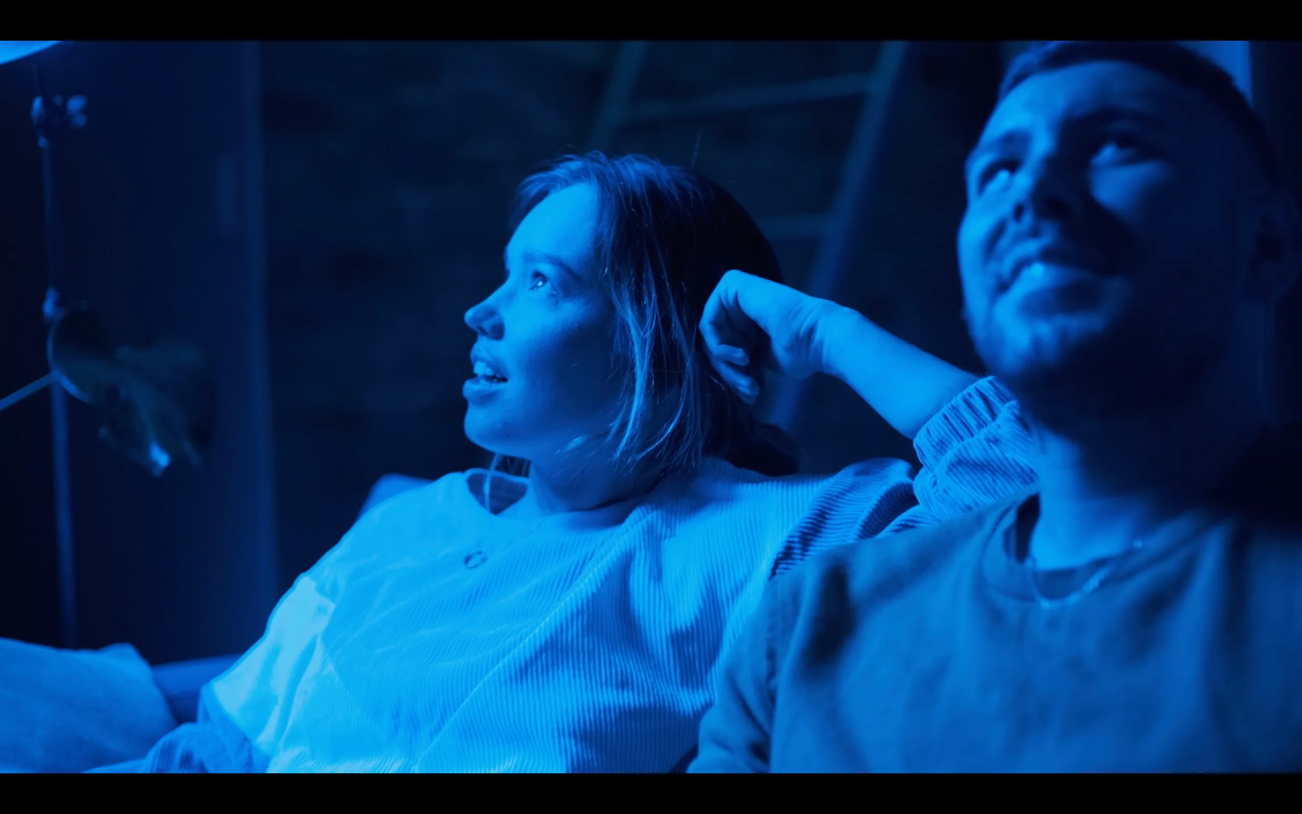 Two people sitting closely on a couch, bathed in blue light, looking upward with relaxed and engaged expressions, suggesting they are watching something together.