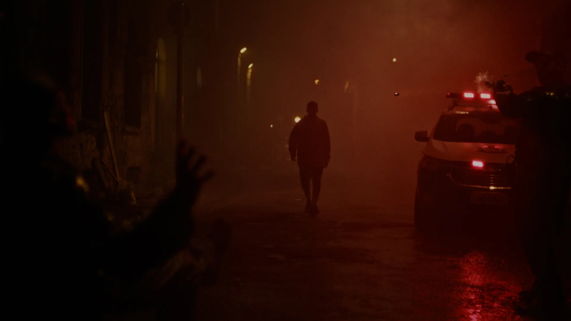 A person walks alone down a dark, foggy street at night. Red and yellow lights from a parked police car illuminate the scene, with two shadowy figures visible in the foreground.
