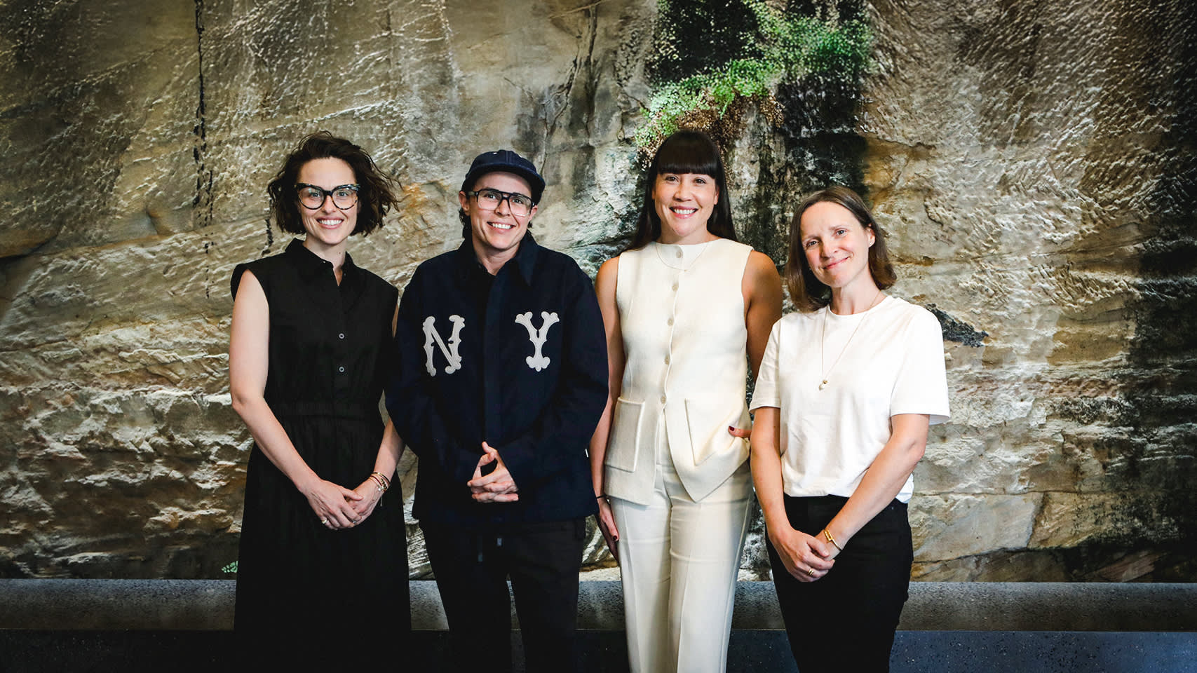 Four people stand side by side, smiling in front of a textured rock wall with greenery. They are dressed in casual and business-casual outfits and facing the camera.