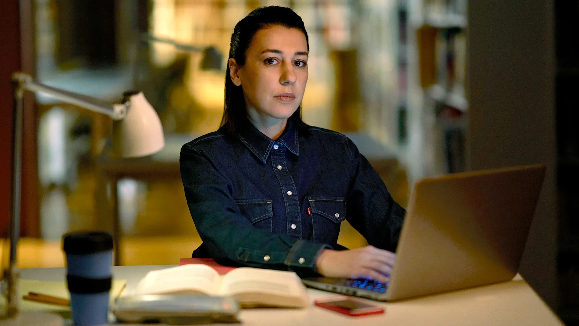 A woman in a denim shirt sits at a desk working on a laptop at night, with an open book, a desk lamp, a coffee cup, and a smartphone in front of her. Bookshelves are visible in the blurred background.
