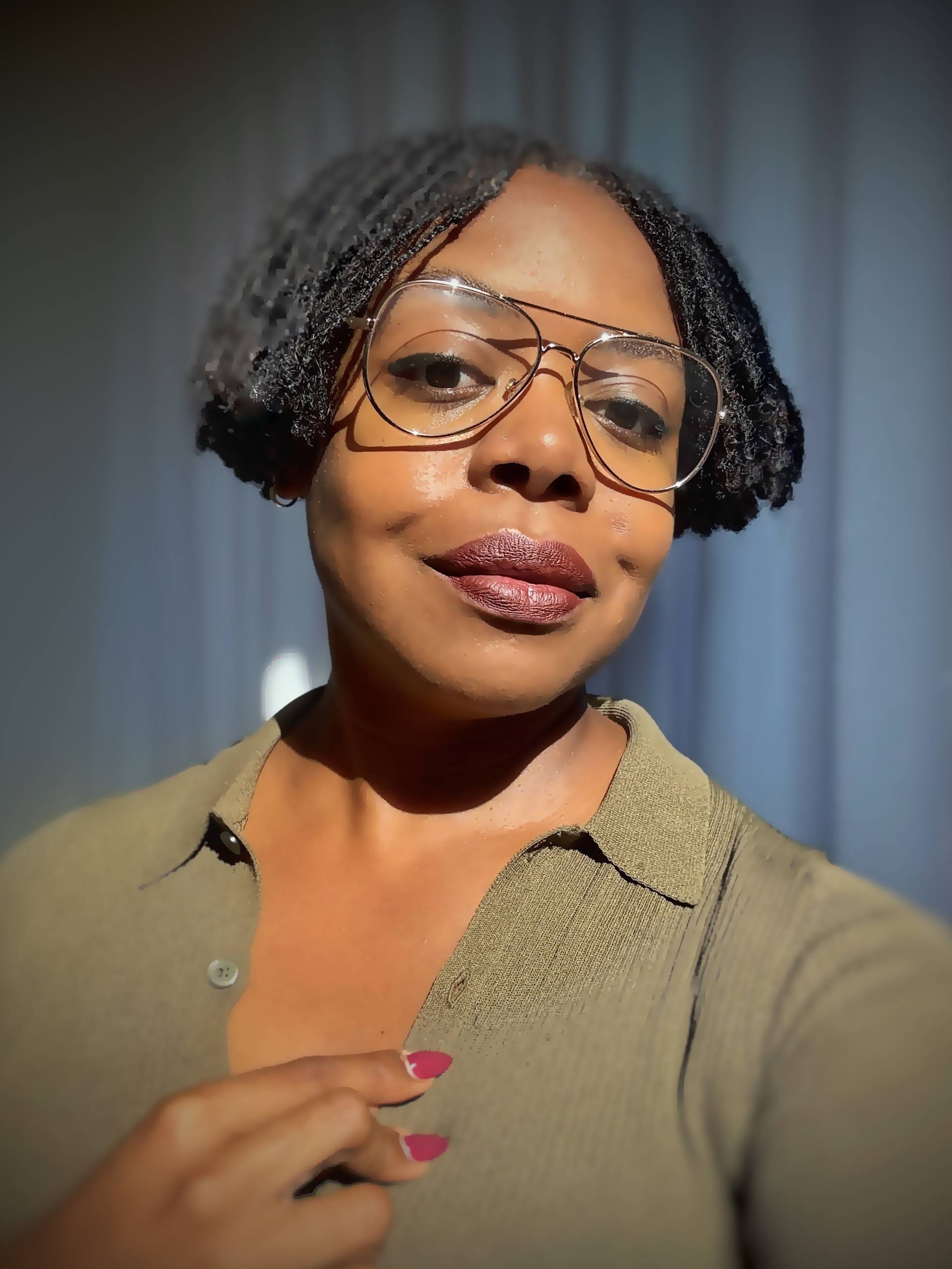 A woman with short, twisted hair wears large glasses and a green collared shirt, smiling slightly. She has dark lipstick and nails painted pink, touching her collar. The background is softly blurred.