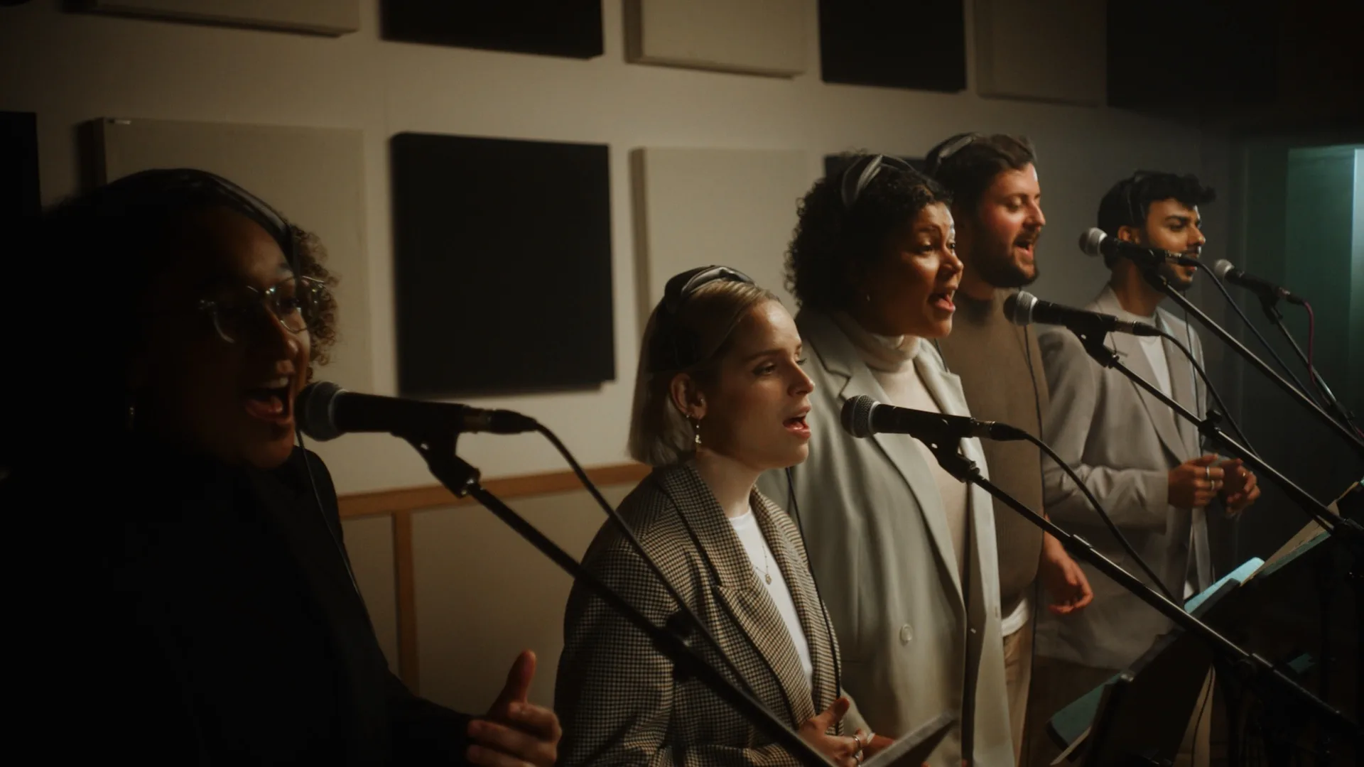 Five people wearing headphones sing into microphones in a recording studio, standing in a row with sound panels on the wall behind them. They appear focused and engaged in their performance.