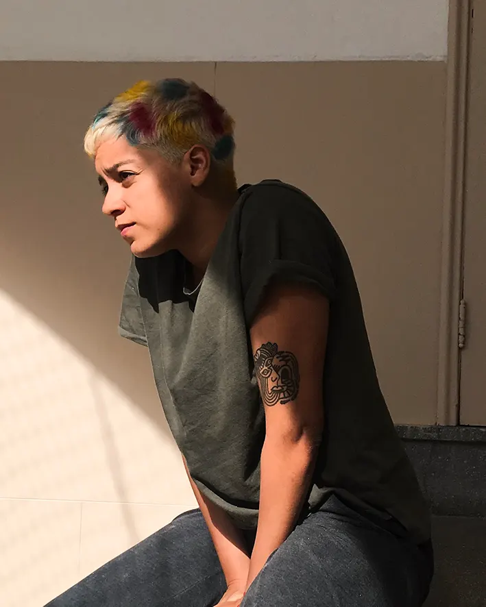 A person with short, multicolored hair sits in sunlight, wearing a dark t-shirt and jeans, with a horse tattoo visible on their upper arm. They look thoughtful, gazing off to the side.