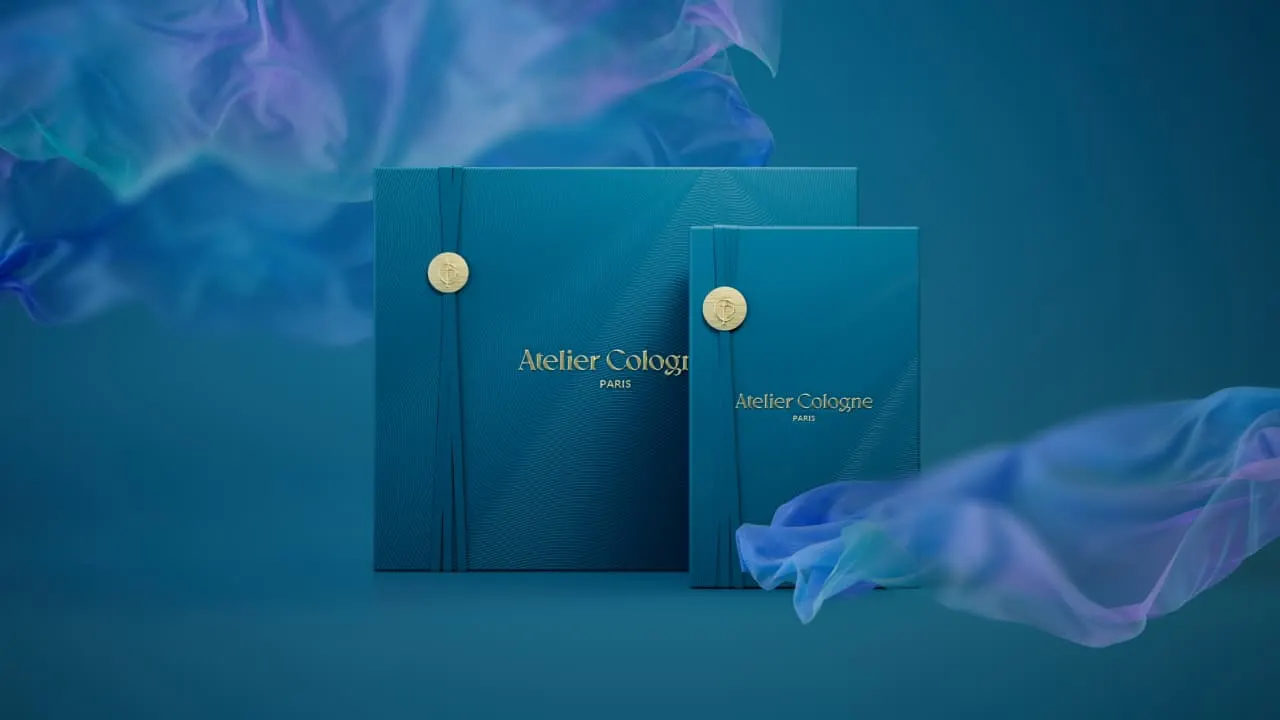 Two elegant, teal-colored boxes with gold seals and “Atelier Cologne Paris” written on the front, set against a blue background with wisps of purple and blue translucent fabric.
