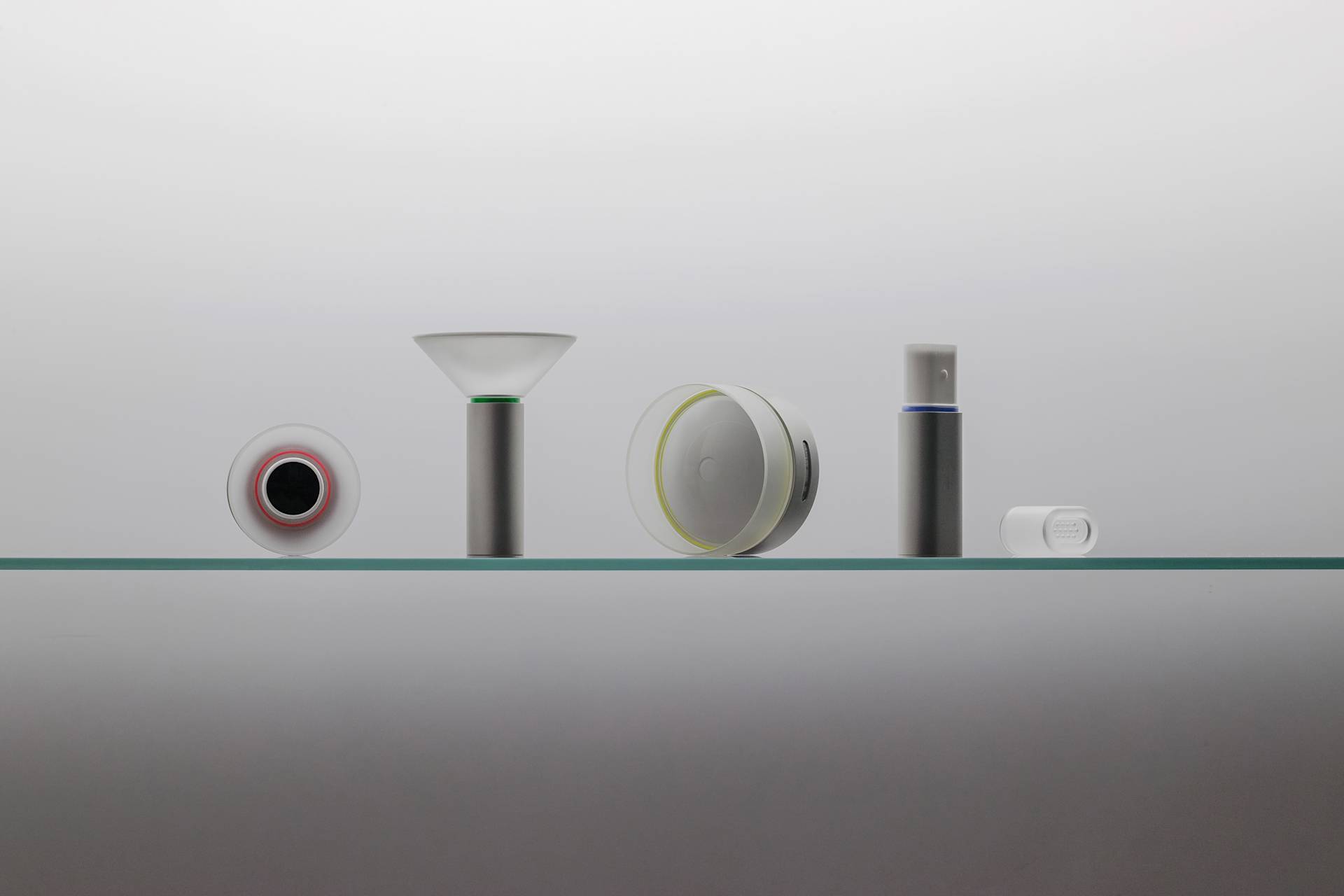 Five minimalistic, modern devices with cylindrical and disc shapes are displayed in a row on a glass shelf against a plain white background. The items are sleek, mostly white or silver, with subtle accent colors.