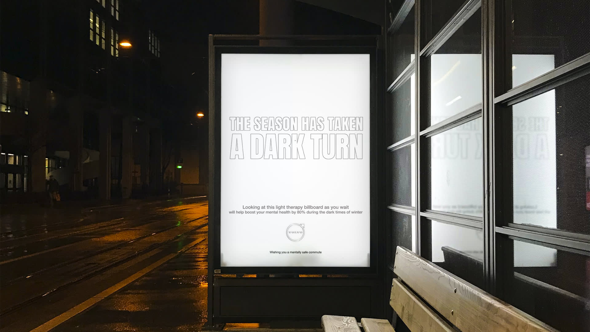 A bus stop billboard at night displays the message The season has taken a dark turn in bold letters on a bright white background, with smaller text below about light therapy for winter mental health. A bench is in front of the sign.