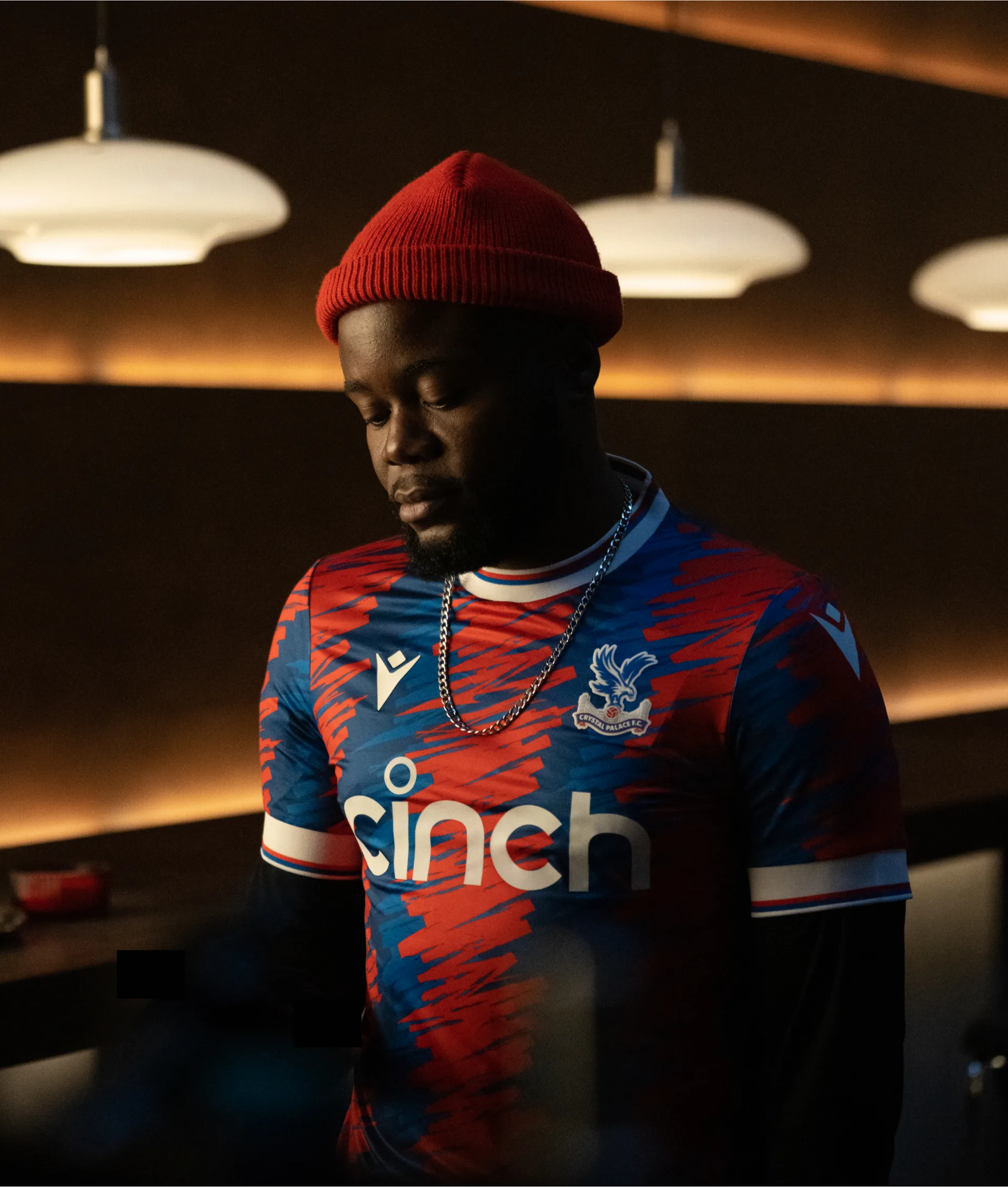 A man wearing a red beanie, a chain necklace, and a colorful Crystal Palace football jersey stands indoors under modern pendant lights, looking down with a thoughtful expression.