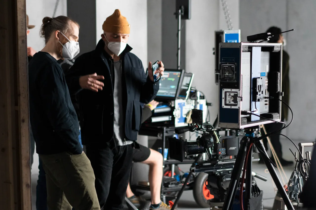 Two people wearing face masks stand and talk on a film set, surrounded by cameras, monitors, and equipment. One gestures with his hands while the other listens. A crew member sits in the background.