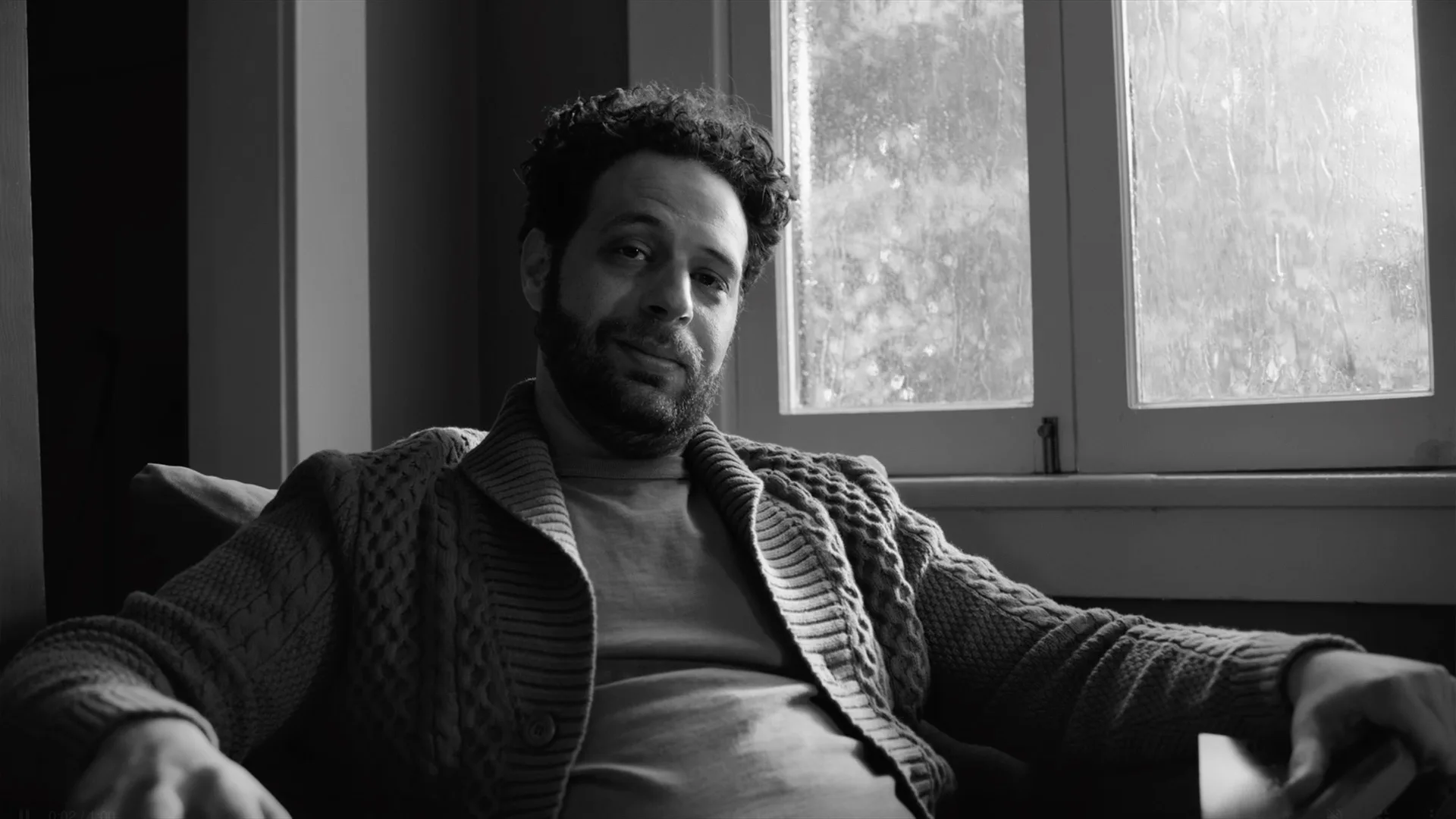 A man with curly hair and a beard sits on a couch near a window, wearing a textured cardigan and t-shirt, looking at the camera with a relaxed expression. The image is in black and white.