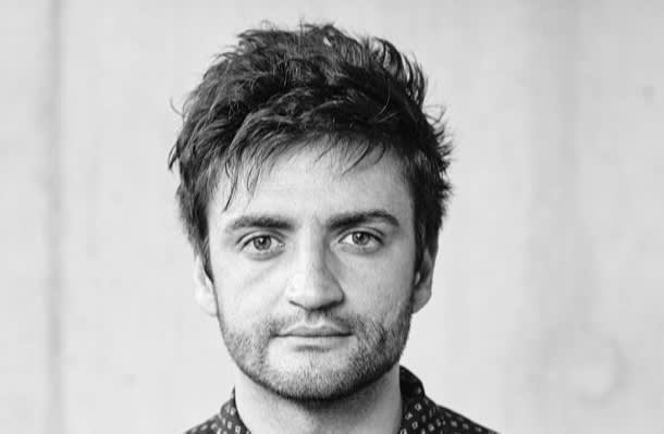 A young man with tousled dark hair and a short beard looks directly at the camera with a neutral expression. The background is plain and out of focus. The image is in black and white.