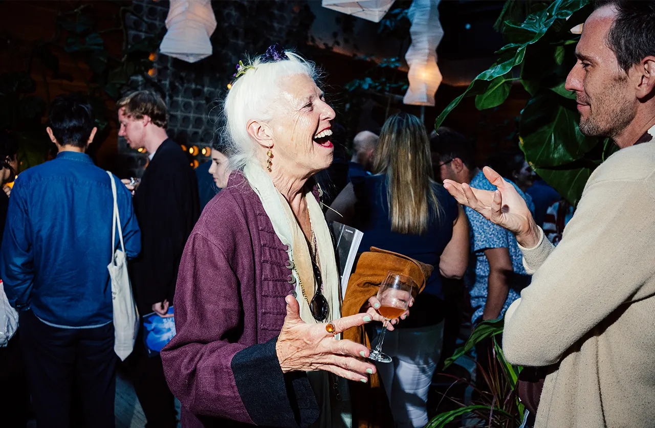 An older woman with white hair laughs and gestures while holding a wine glass at a lively indoor event, surrounded by people engaged in conversation.