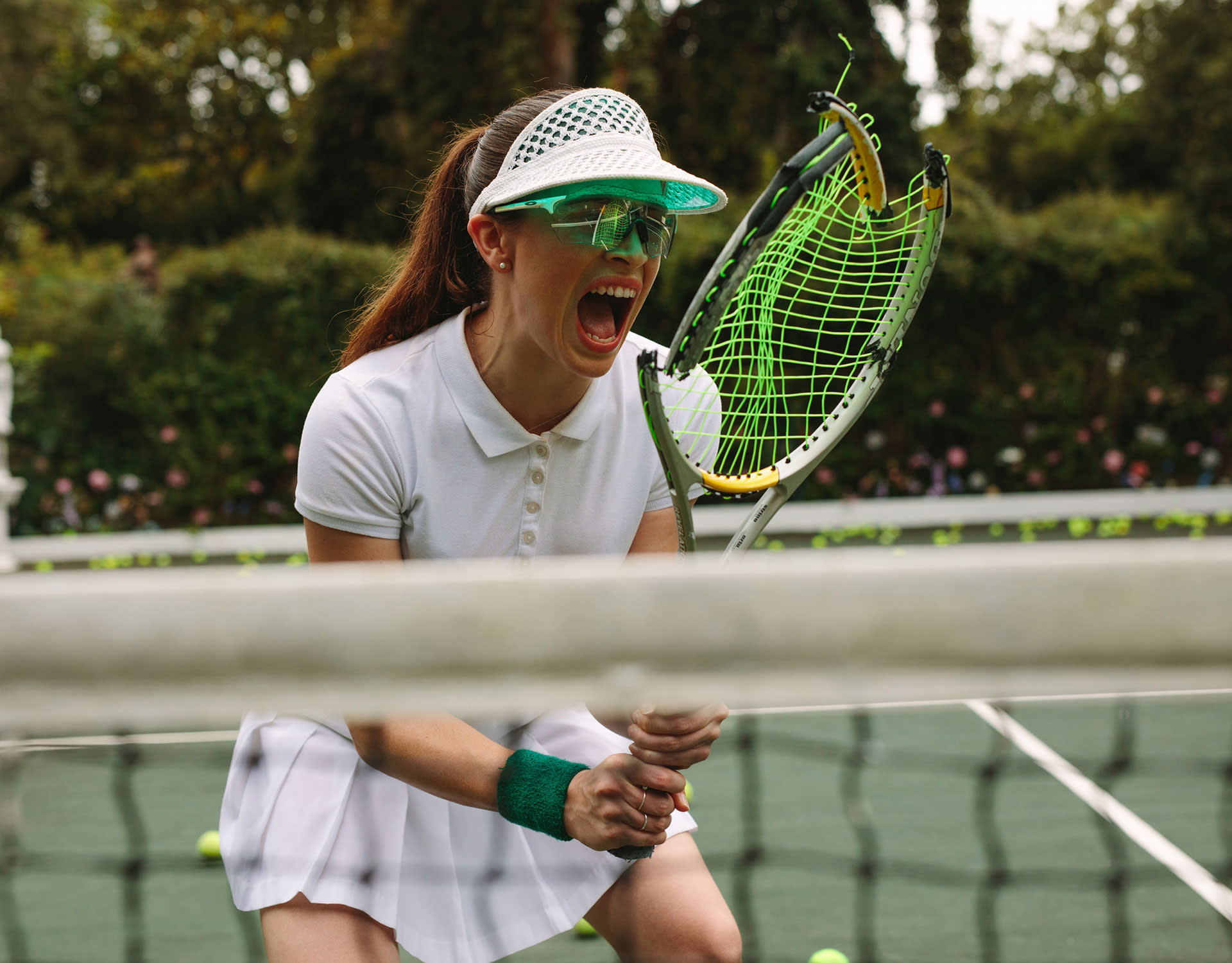 A tennis player in white sportswear shouts in frustration on the court, holding a broken racket with bent strings and frame. Tennis balls are scattered on the ground in the background.